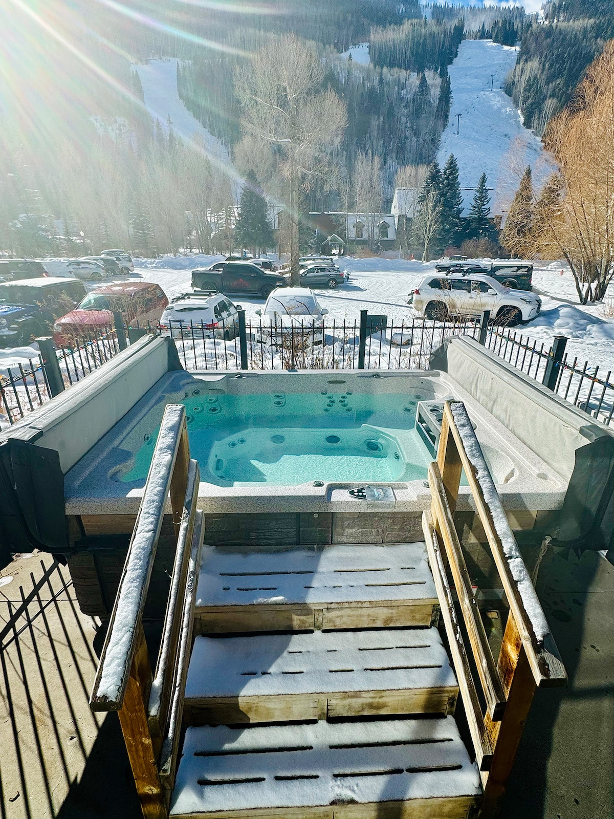 An outdoor hot tub is positioned on a deck with wooden steps leading down. Scenic mountain slopes are visible in the background, flanked by trees. Sunlight illuminates the area, reflecting off the water's surface, creating a serene environment.