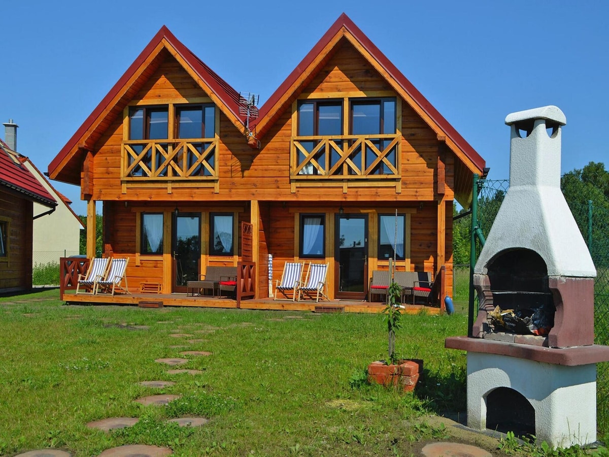 A semi-detached wooden house features a red roof and two stories, framed by large windows. A small outdoor fireplace is present in the foreground, complemented by seating areas and a grassy lawn with stepping stones leading towards the entrance.