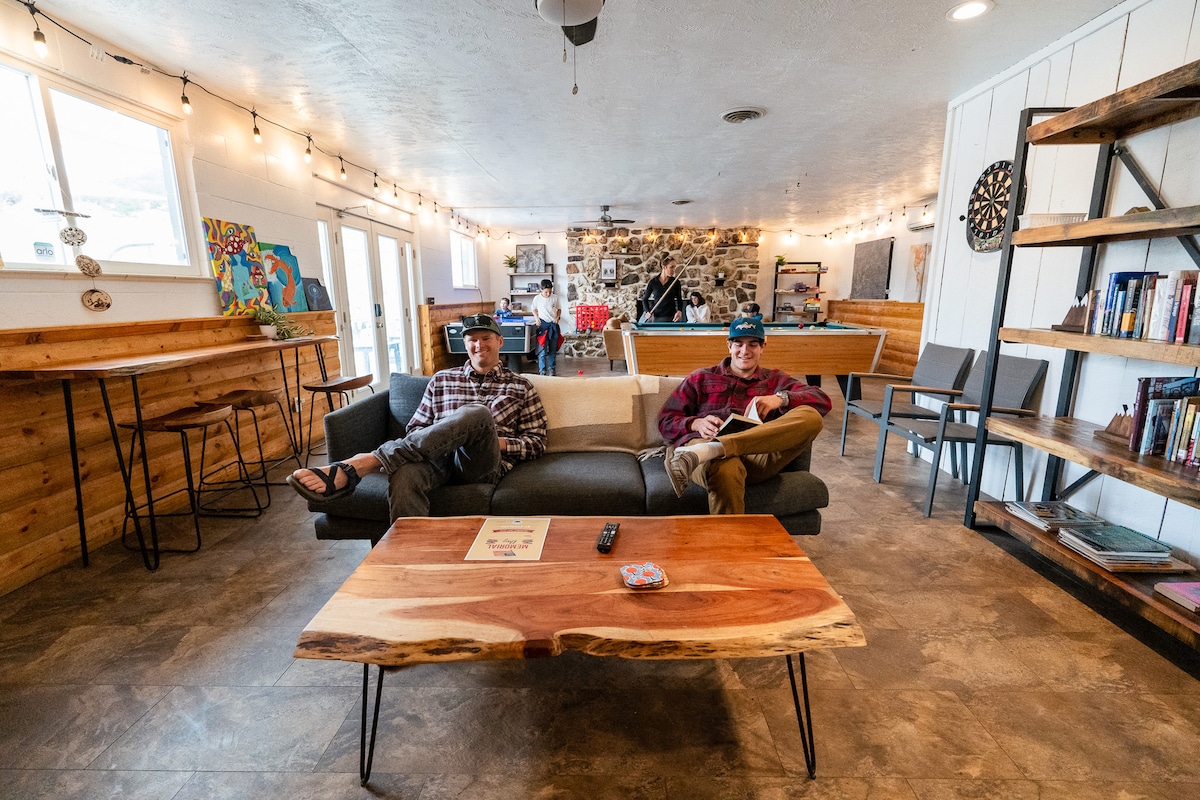 A spacious communal lounge features a wooden table and a comfortable sofa. Two individuals sit relaxed, engaging with their devices. Large windows provide natural light, while a stone wall adds to the rustic decor. Various seating arrangements and a dartboard are visible in the background.
