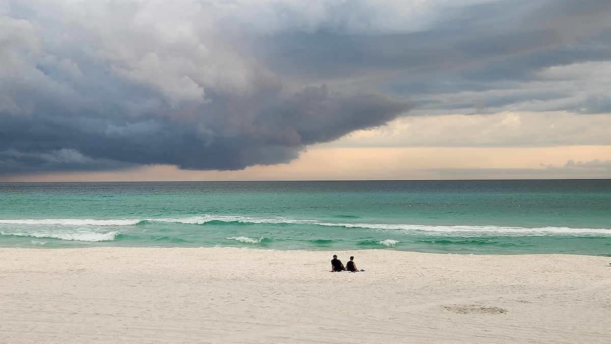 A serene beach scene features two individuals seated on the white sandy shore, gazing at the emerald sea. Clouds gather above the horizon, casting soft shadows on the water, while gentle waves lap the shore, creating a tranquil ambiance.