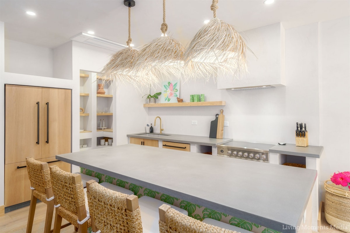 A modern kitchen features a large island topped with a grey countertop, accompanied by four woven bar stools. Unique pendant lights with a natural look hang above. Open shelving displays tropical decor, while wood cabinetry and stainless steel appliances create a clean and inviting space.