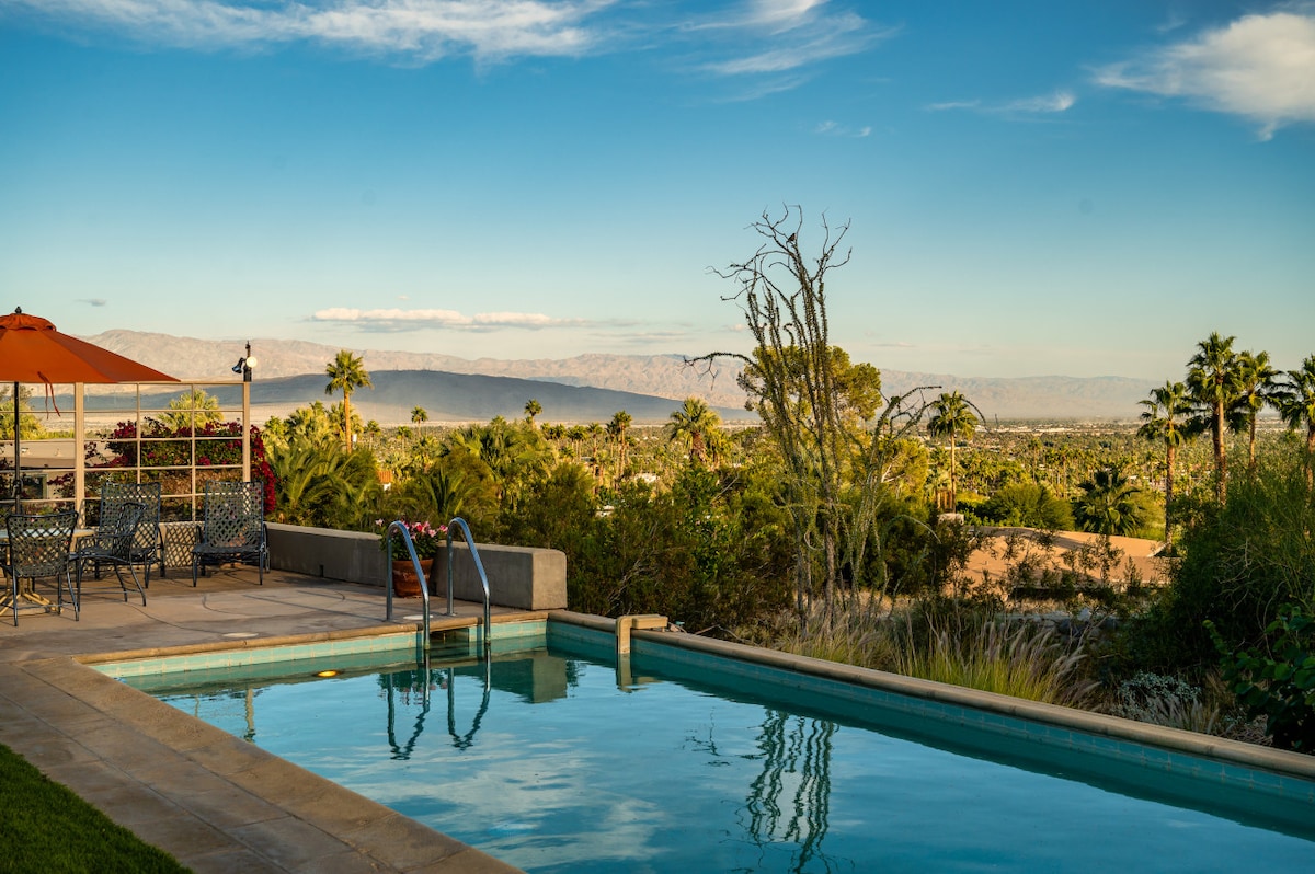A private pool glistens under the sun, offering a serene view of surrounding palm trees and distant mountains. Lounge chairs and an umbrella are arranged nearby, inviting relaxation. The sky is clear, enhancing the tranquil outdoor atmosphere.