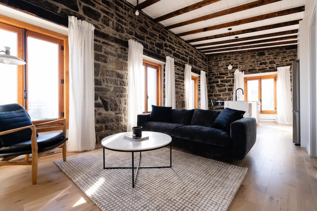 A light-filled living area features exposed stone walls and wooden beams that complement modern furnishings. A black sofa is paired with a round coffee table, while a comfortable chair provides additional seating. Large windows draped with sheer curtains enhance the spacious ambiance.