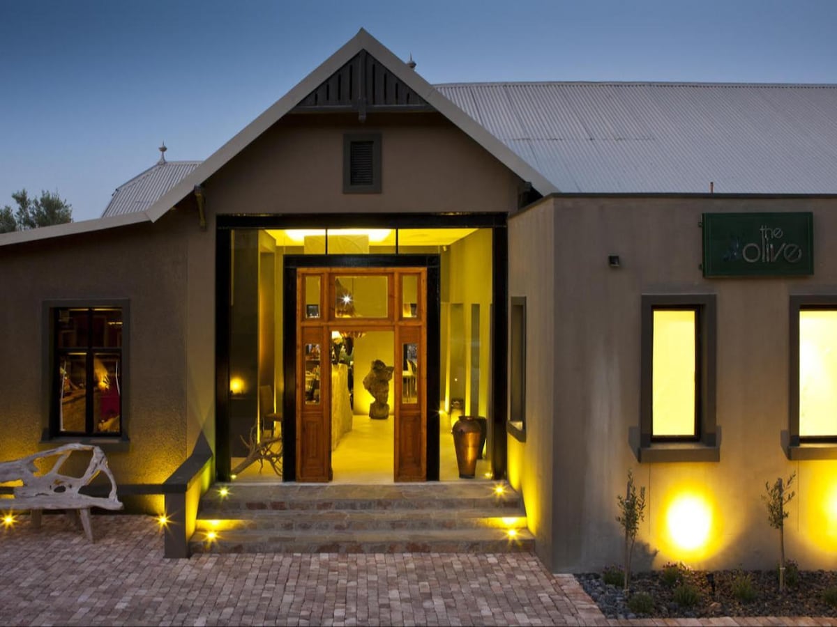 The entrance of the building showcases double wooden doors framed by large glass panels. Soft lighting highlights the steps leading to the entry, and decorative elements such as a sculpture and potted plants provide a welcoming touch. Side windows create a bright atmosphere.
