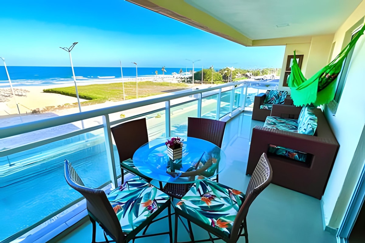 A spacious balcony features a circular glass table surrounded by four textured chairs. Colorful decorative cushions add comfort to a nearby seating area. A hammock is draped to one side, and the beautiful ocean view is visible beyond the glass railing.