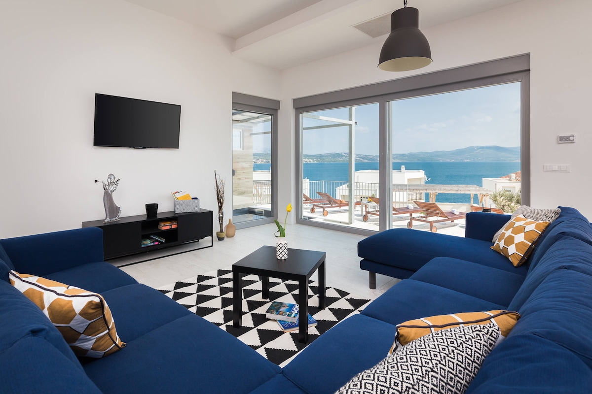A modern living area is highlighted by a large blue sectional sofa, facing a wall-mounted flat-screen TV. Floor-to-ceiling windows provide a panoramic sea view and ample natural light, while a black and white patterned rug adds contrast to the light-colored tiles.