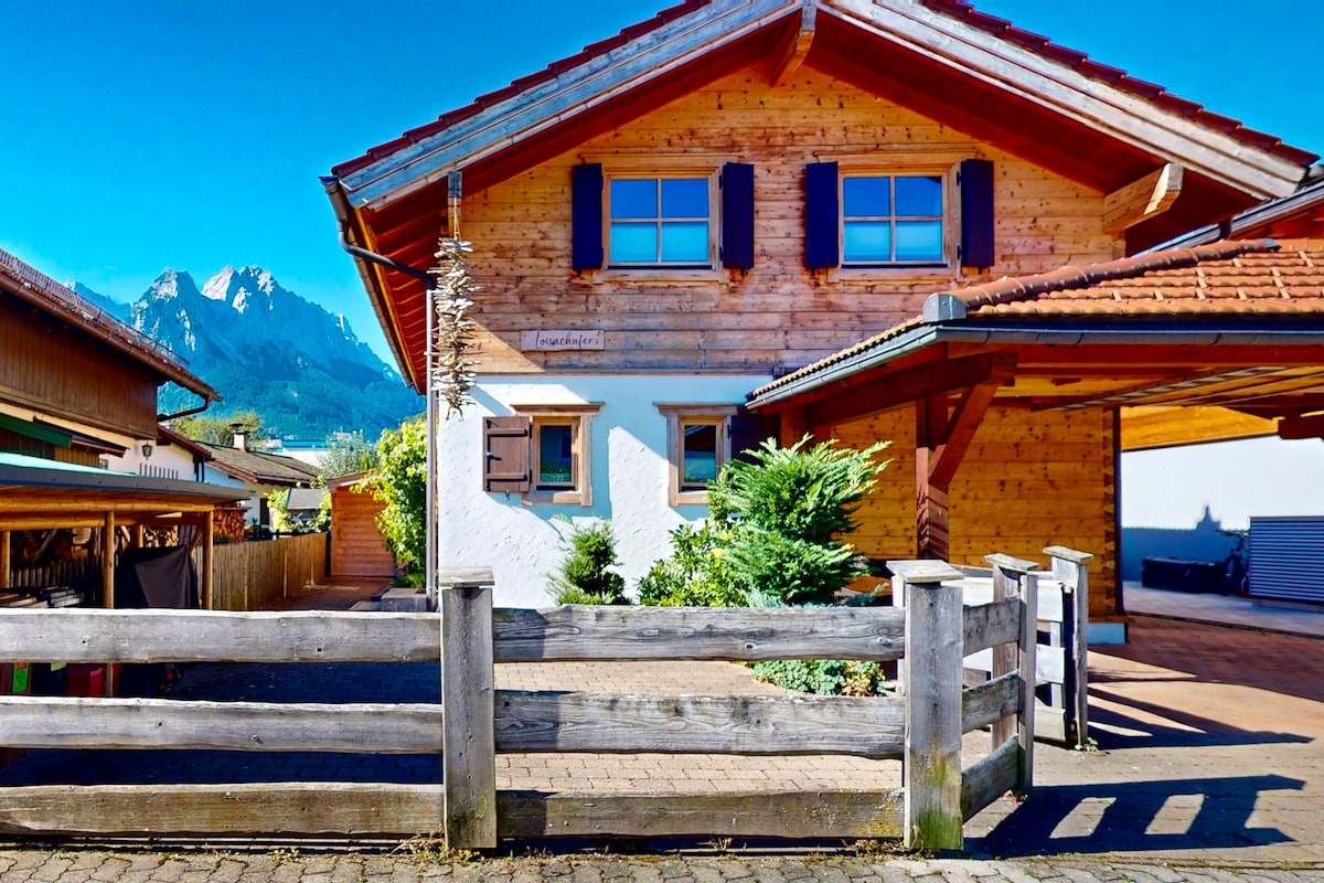 A wooden cottage is presented with a combination of stone and sloping roof. The exterior features large windows and traditional shutters. A fenced yard surrounds the property, with mountains visible in the background against a clear blue sky.