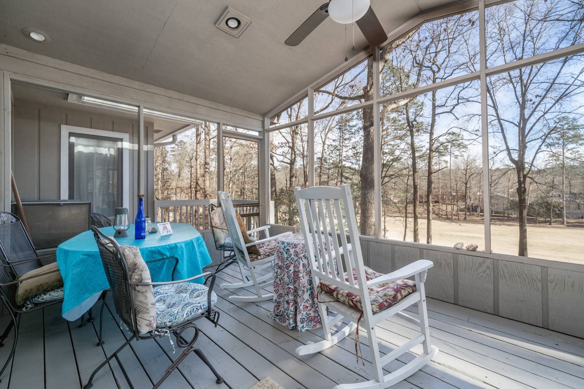 A screened-in porch features rocking chairs and a large dining table set with a blue cloth. The surrounding area offers expansive views of nature, with trees visible in the background. Sunlight filters through the screens, creating a bright and airy space for relaxation.