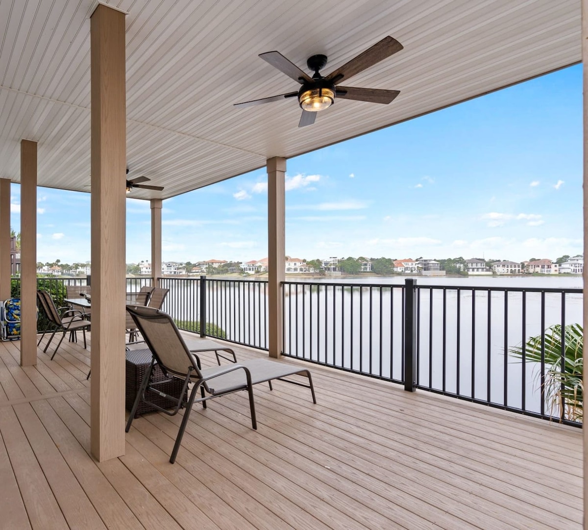 A spacious balcony features comfortable lounge chairs with views of a serene lake. The area is shaded by a covered ceiling, and ceiling fans are installed for added comfort. The smooth wooden flooring complements the railing that borders the balcony, enhancing the outdoor experience.