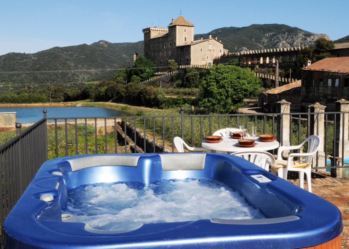 An outdoor jacuzzi is positioned on a patio, with views of a castle and rolling hills in the background. A dining table with chairs is nearby, providing an area for outdoor meals. The clear blue sky enhances the serene setting.
