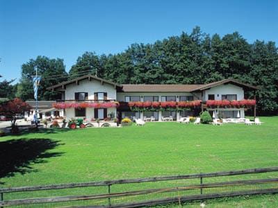 The guest house features a two-story structure with a welcoming facade adorned with colorful flowers. A large, well-maintained lawn surrounds the building, providing ample space for relaxation. The backdrop includes a dense forest, enhancing the tranquil setting.