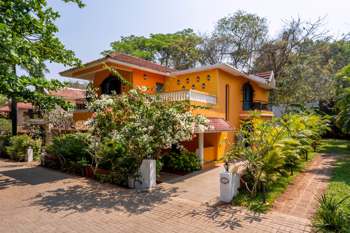 The villa features a vibrant yellow exterior, framed by lush greenery and blooming flowers. A spacious driveway leads to the entrance, while multiple levels of the structure add dimension. Balconies adorn the upper floor, allowing for outdoor relaxation.