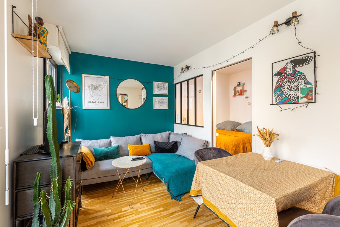 An open-plan living room features a cozy corner sofa in gray with blue and yellow throw blankets. A round table is accompanied by two chairs, and a large mirror reflects natural light. Plants add a touch of greenery, enhancing the overall ambiance.