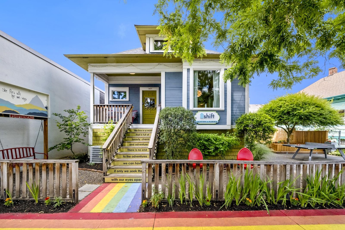 The entrance to the duplex is marked by colorful steps leading to a welcoming porch. A vibrant garden with various plants and trees frames the entrance, while two red chairs accompany a wooden bench, inviting guests to relax outdoors.