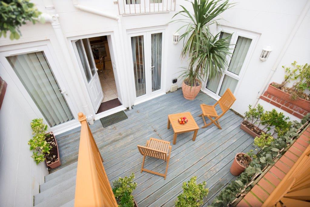 A private decked area features wooden furniture, including two chairs and a table, surrounded by potted greenery. Natural light filters in through nearby glass doors, leading to the apartment's interior, enhancing the inviting outdoor space.