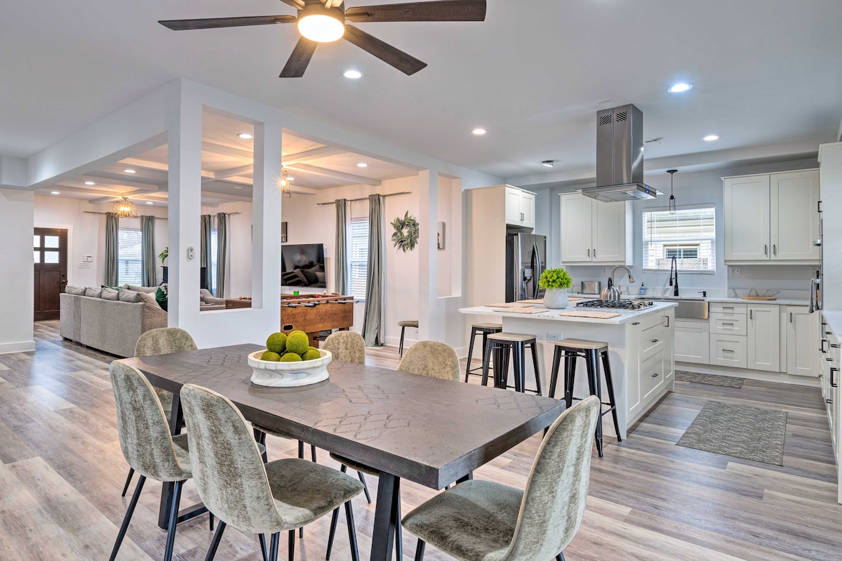 A modern kitchen and dining area feature a spacious table surrounded by soft upholstered chairs. Bright cabinetry and stainless steel appliances are visible, and overhead lighting illuminates the space. A large island with seating is positioned centrally, while floor-to-ceiling windows provide natural light.