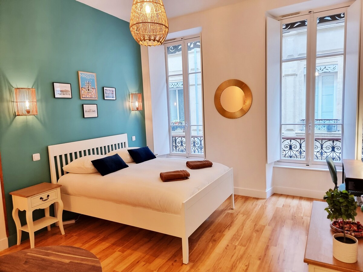 A cozy sleeping area features a spacious bed with two rolled towels placed neatly atop. Soft blue-green walls complement decorative art pieces. Natural light enters through two large windows, framed with elegant sheer curtains, illuminating the light wooden floor and a small side table.