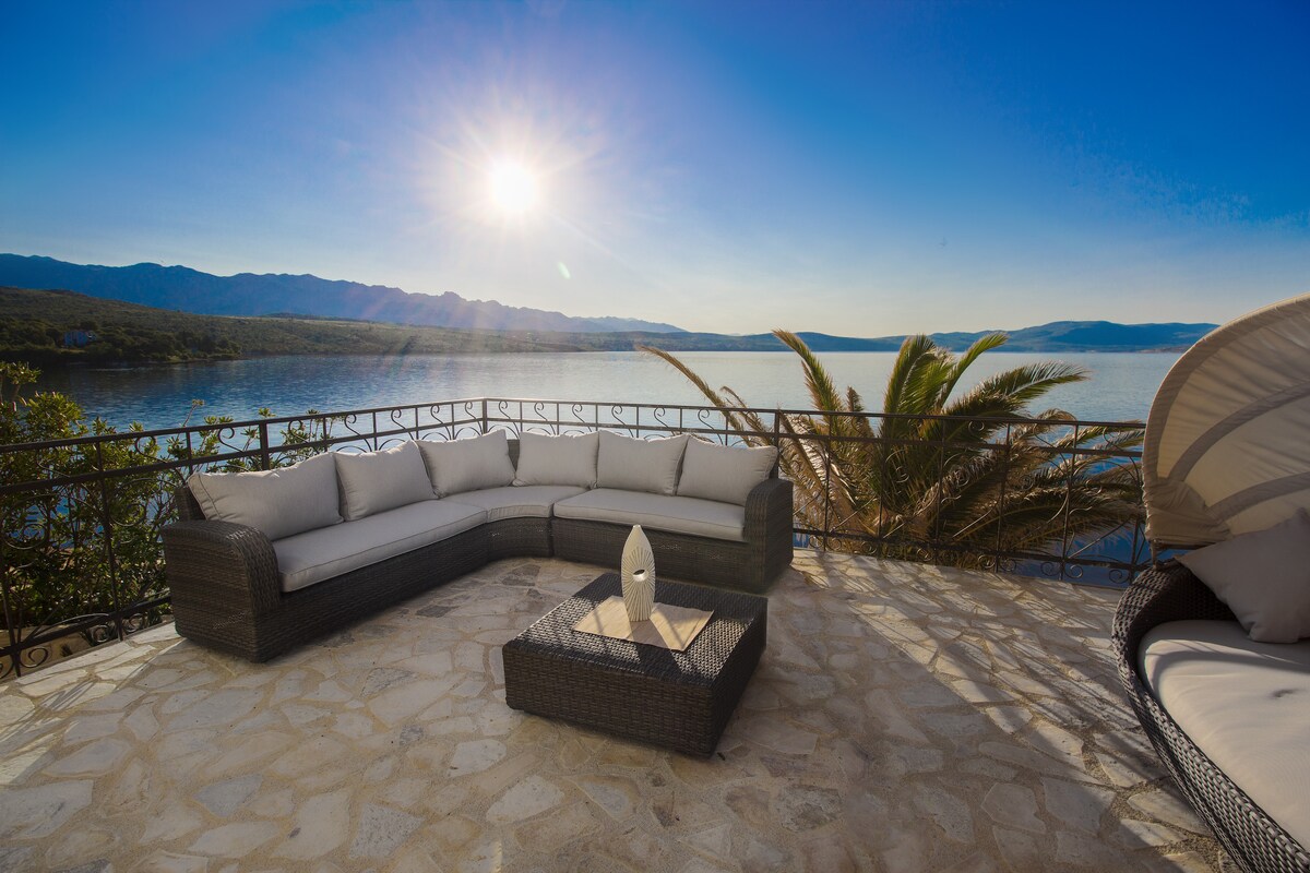 A spacious outdoor seating area features a curved sectional sofa with soft cushions, positioned on a stone terrace. A coffee table is placed in the center, surrounded by scenic views of the water and mountains, with sunlight illuminating the scene.