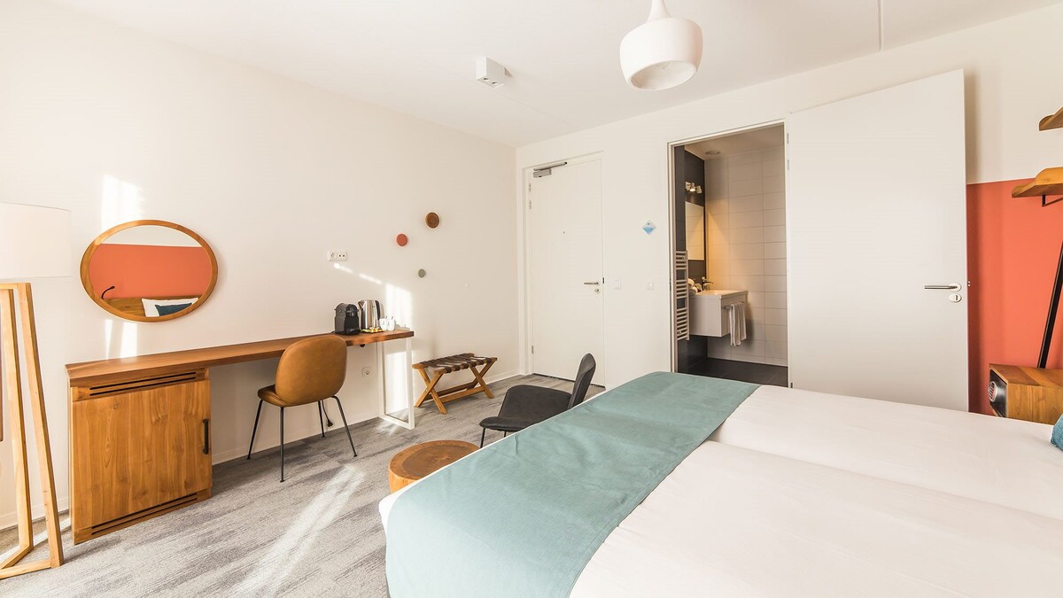 A bright hotel room is featured, showcasing twin beds with light blue bedding. A work desk with a chair and a mirror is positioned near a window, while a seating area includes a wooden stool. A door leads to a bathroom, partially visible in the background.