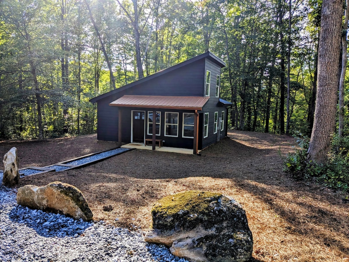 The modern exterior of the retreat is framed by trees, offering a sense of privacy. A sloped roof and large windows enhance natural light. A pathway leads to the entrance, while a gravel area features round stones that add texture to the landscape.
