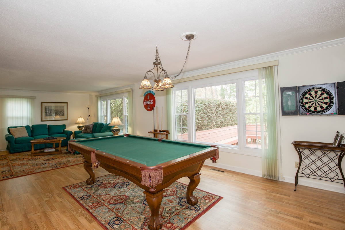 A spacious game room features a green felt billiard table at its center, surrounded by light hardwood floors. Large windows allow natural light to fill the room, showcasing comfortable seating and a dartboard nearby. Decorative elements, including a Coca-Cola sign, add a casual touch.