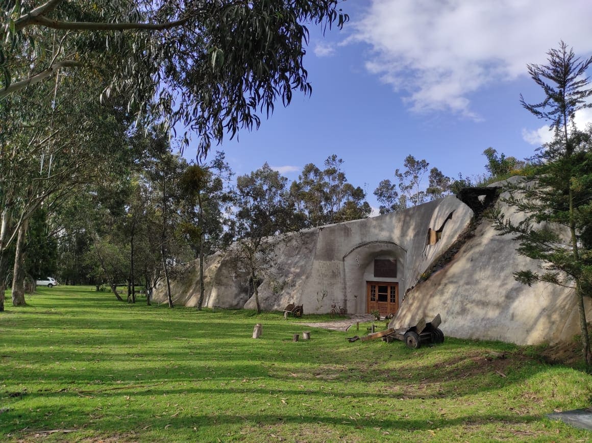 A natural setting features a unique cave-like exterior nestled within a grassy area, surrounded by tall eucalyptus trees. The entrance is framed by greenery, with a glimpse of wooden doors visible, suggesting an inviting entry into the cave.