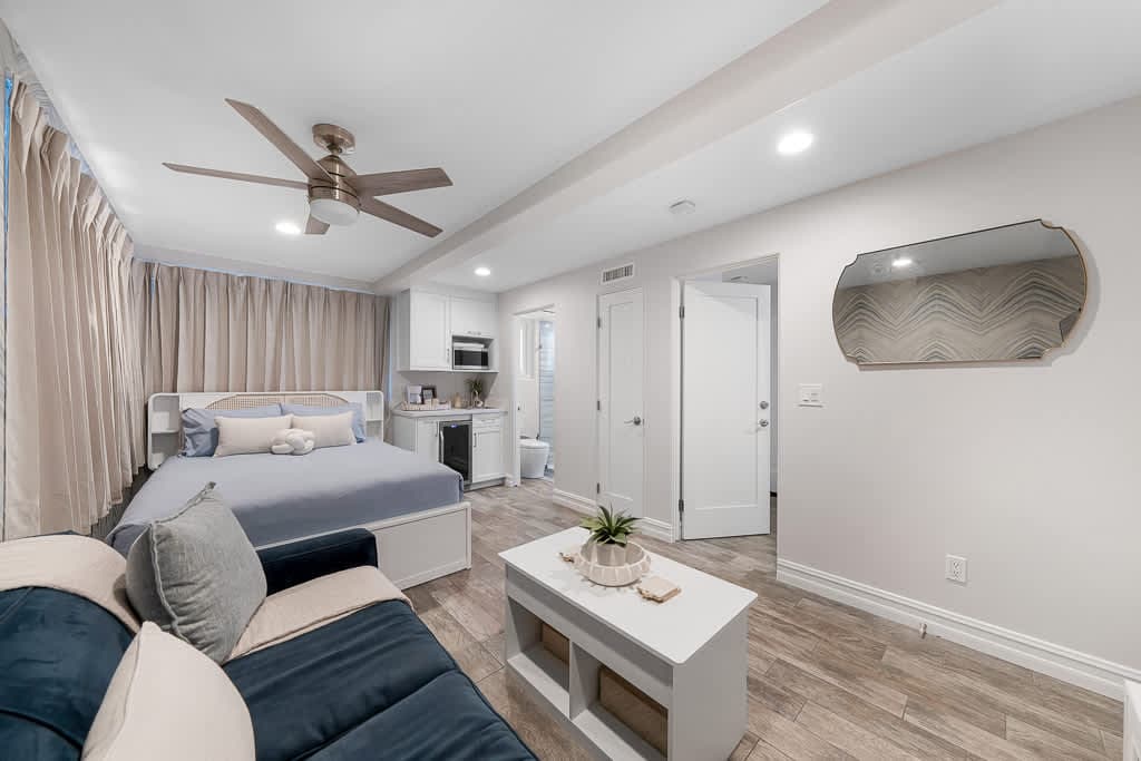 A spacious studio is featured, showcasing a queen bed with a light blue coverlet and neutral-toned furnishings. A comfortable sofa is present, accompanied by a stylish coffee table. Light-colored cabinetry and appliances are visible in the kitchenette area. A ceiling fan and soft lighting enhance the ambiance.