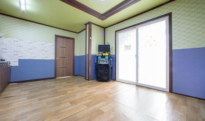 The spacious interior features light wood flooring and contrasting blue and green walls. A door allows natural light to enter, while a small entertainment area with a television and audio system is present against one wall.