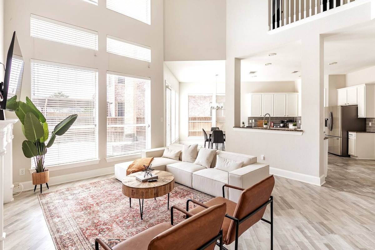 An open living area features large windows allowing natural light to fill the space. A modern sectional sofa sits adjacent to a round wooden coffee table, while two leather chairs provide additional seating. The minimalist design is complemented by a warm area rug on the light-colored flooring.