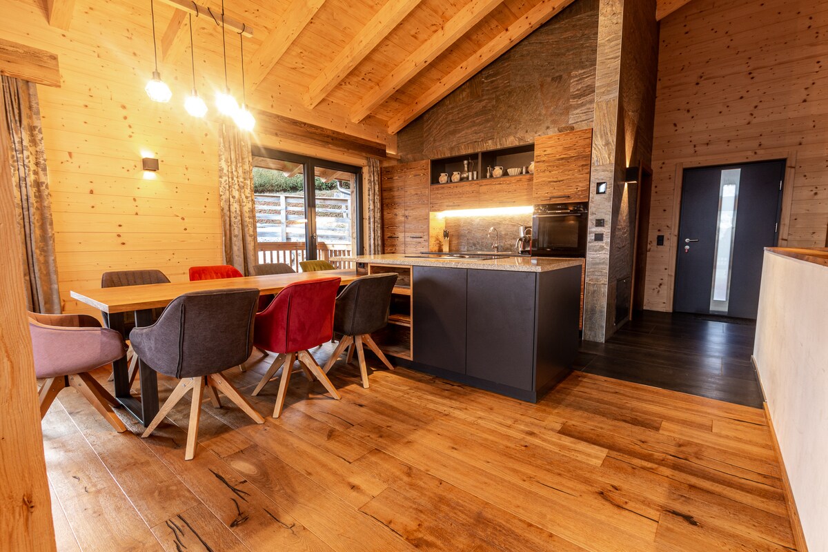 The open kitchen-living room area features a large dining table surrounded by six colorful chairs. A modern kitchen island with appliances is set against a wooden backdrop, and large windows provide views of the outside patio. Warm wooden beams enhance the space's inviting atmosphere.