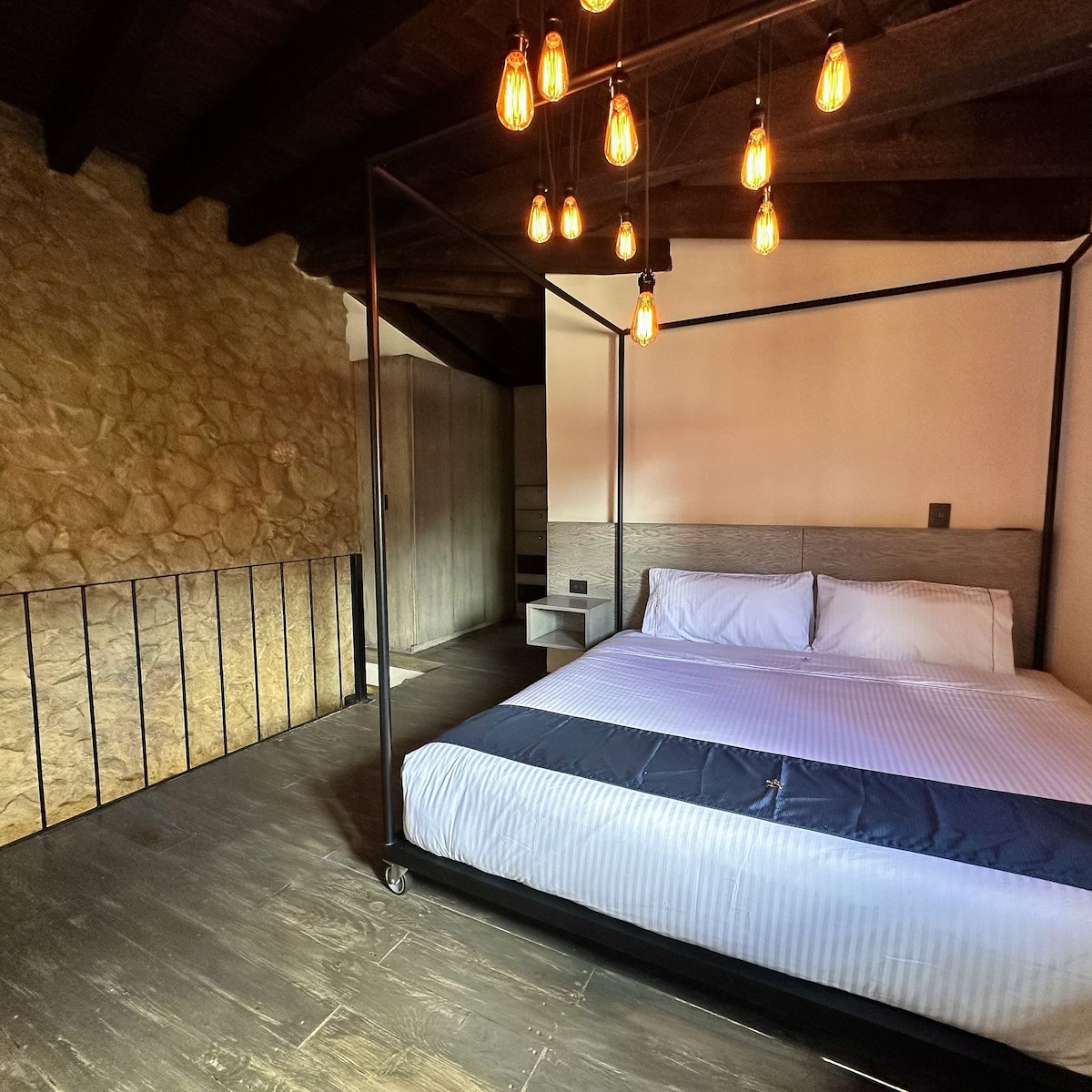 A spacious sleeping area is defined by a four-poster bed with crisp white linens. Soft lighting is provided by hanging Edison bulbs above the bed, while textured stone and wooden elements accentuate the rustic charm of the loft.
