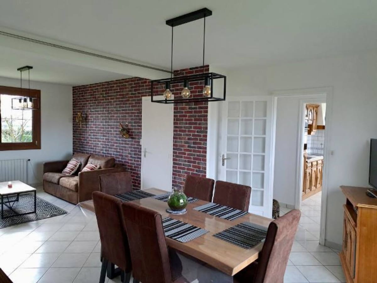 A spacious living and dining area features a brick accent wall and a modern pendant light. A wooden dining table with dark chairs is set for meals, while a comfortable sofa offers seating for relaxation. Large windows invite natural light into the room.