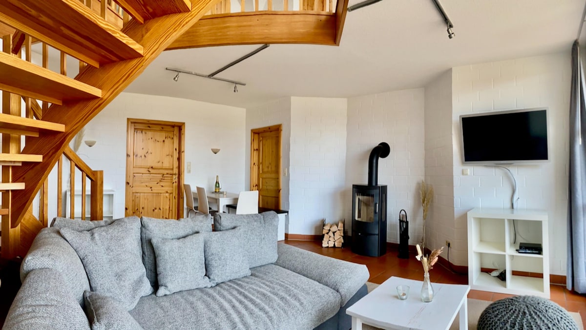 A spacious living area features a gray sofa with several cushions, positioned near a modern wood stove. The wooden staircase leads to the upper floor, while two wooden doors and a television mounted on the wall are visible in the background.