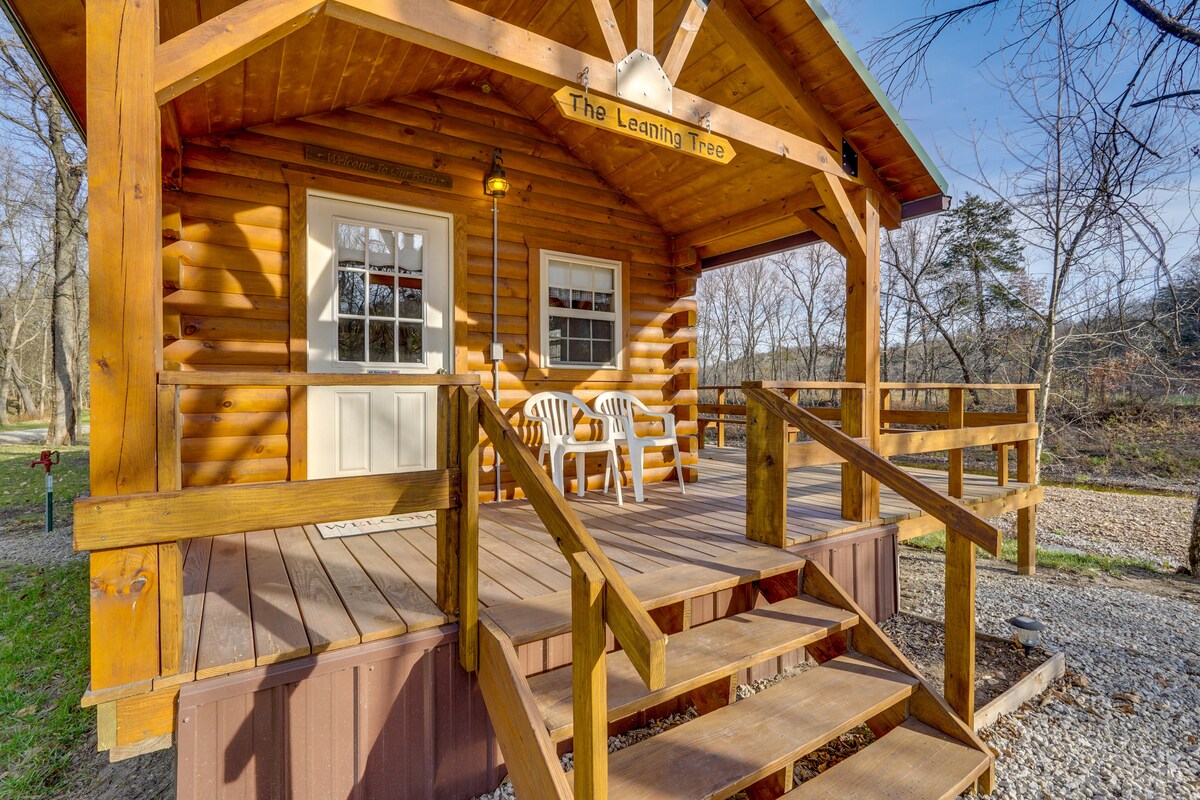 The rustic exterior of the cabin features wooden logs and a welcoming front porch with two white chairs. Stairs lead up to the entry, which is framed by windows that allow natural light to fill the space. Surrounding views of trees and gravel paths are visible.