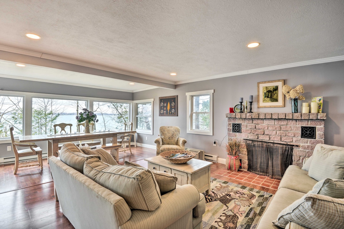 A spacious living room features large windows that offer views of Sawmill Bay. Comfortable seating arrangements include a soft sofa and armchairs, while a rustic fireplace adds character to the space. A dining table is positioned nearby, enhancing the room's inviting ambiance.