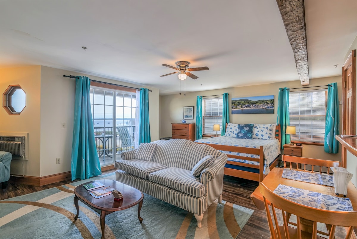 The studio apartment features a spacious layout with a queen-size bed and a comfortable sofa. Large windows allow natural light, while the private balcony offers views of the beach. A dining area is present, complemented by a decorative rug and wooden furnishings.