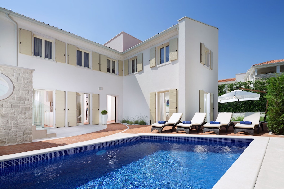 A bright and airy exterior of the villa is showcased, featuring light-colored walls and decorative shutters. Sun loungers are arranged by the refreshing pool, while a patio umbrella provides shade. The inviting pool reflects the clear blue sky, enhancing the serene atmosphere of the outdoor space.