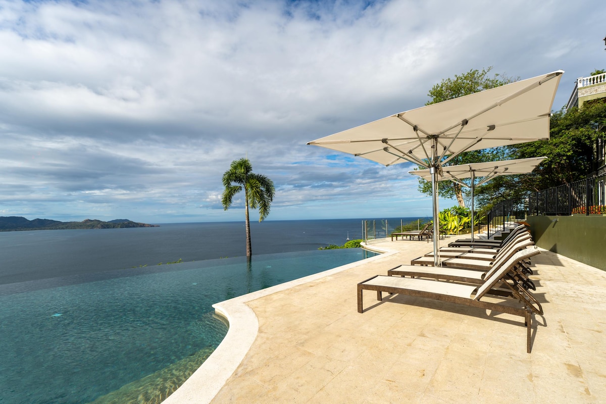 An infinity salt-water pool extends towards the ocean, offering panoramic views of the coastline. Sun loungers are arranged beside the pool, shaded by large umbrellas. Lush greenery surrounds the area, creating a tranquil atmosphere with the sky reflecting in the water.