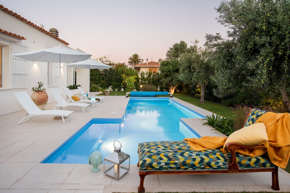 A tranquil outdoor area is highlighted by a clear blue swimming pool, surrounded by sun loungers and umbrellas. A cozy chaise lounge, adorned with bright pillows, sits next to the pool. Lush greenery and decorative lanterns create a calming atmosphere.