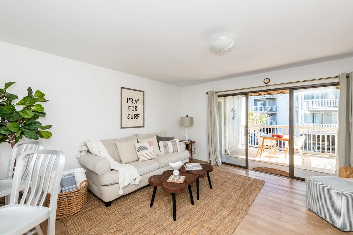 A light and airy living area features a soft, neutral sofa with decorative pillows. Light wood flooring enhances the room's spacious feel. A sliding glass door opens to a balcony, allowing natural light to flood the space. A round jute rug adds texture under a low coffee table.
