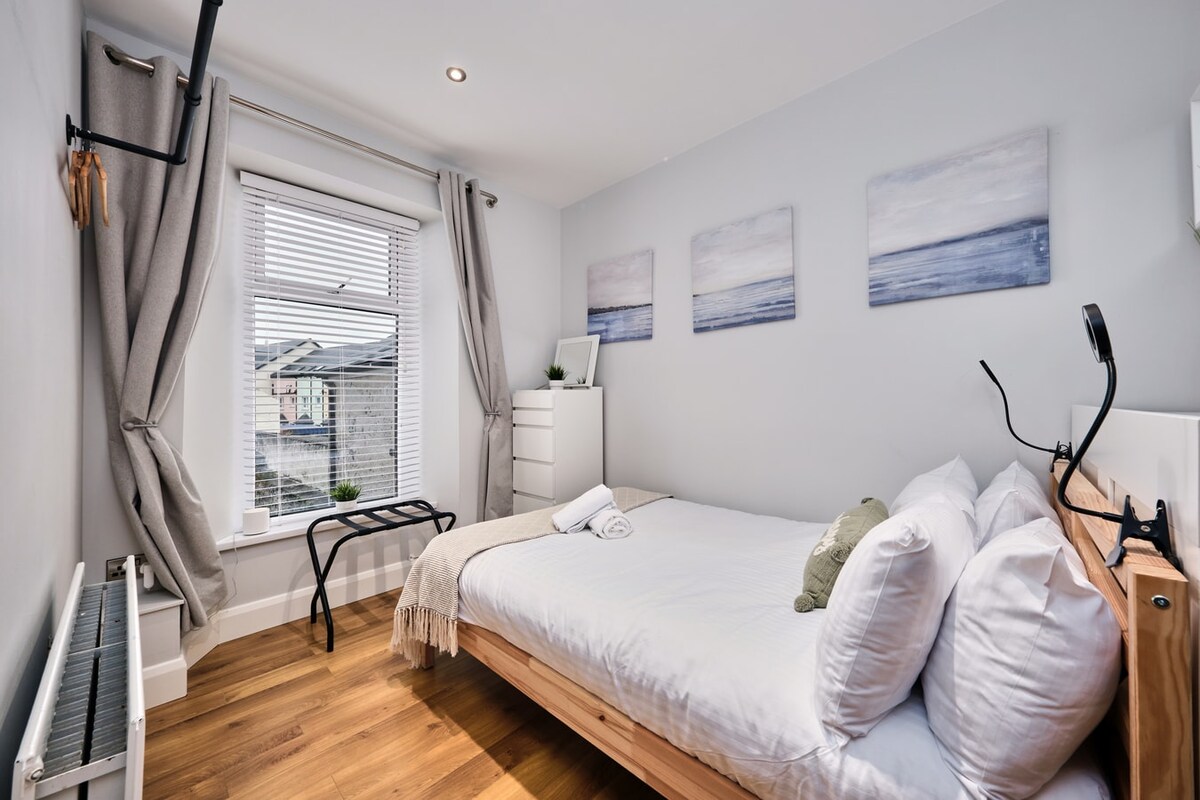A comfortable bedroom is displayed with a spacious bed dressed in crisp linens and pillows. Light streams in through a window adorned with sheer curtains. Modern decor includes three ocean-themed artworks and minimalistic furniture, contributing to a calming atmosphere.