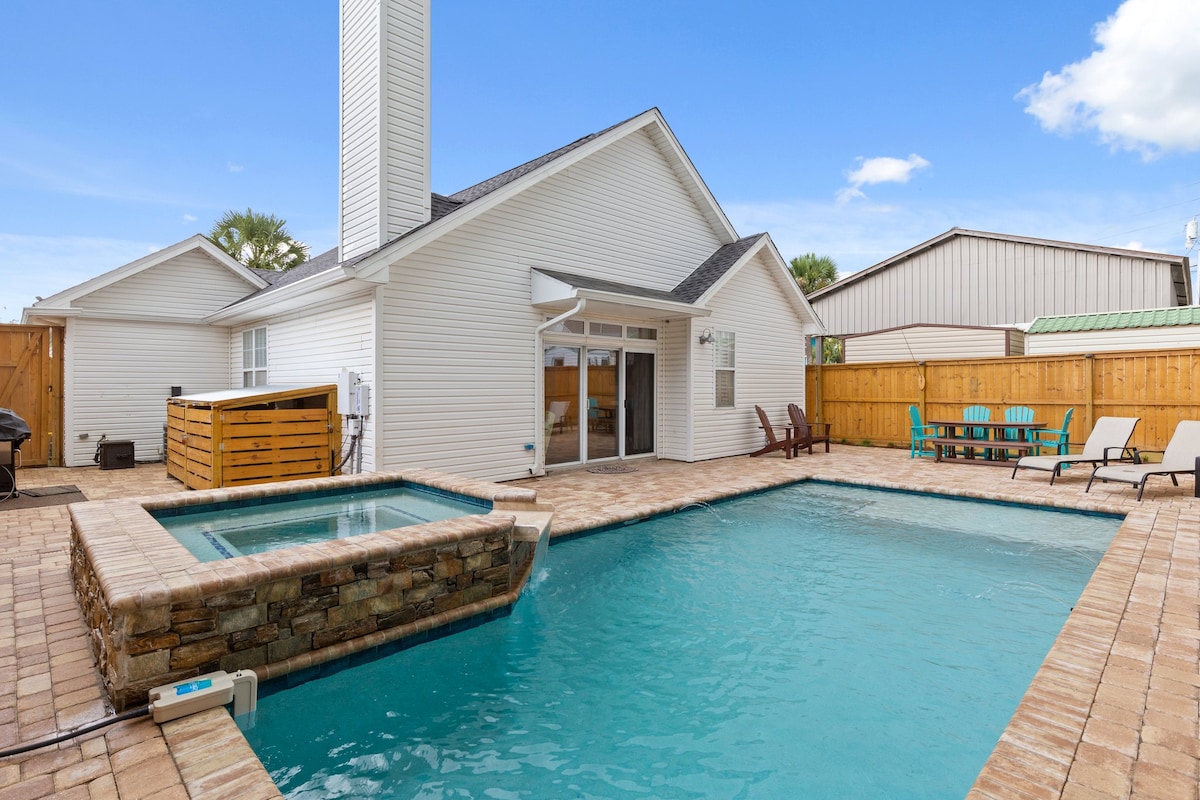 An expansive outdoor area features a private pool and spa, surrounded by stone accents. Lounge chairs and Adirondack chairs are positioned for relaxation. A wooden fence adds privacy, and clear blue skies complement the inviting ambiance of the space.