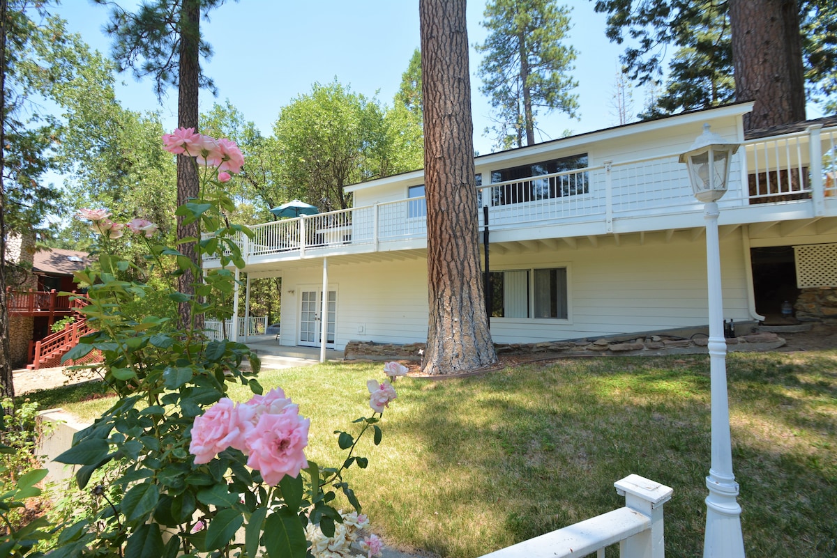 The exterior of a two-story home is presented, surrounded by lush greenery and tall trees. A large deck with a railing extends from the upper level, while pink roses bloom in the foreground, adding a touch of color to the outdoor space.