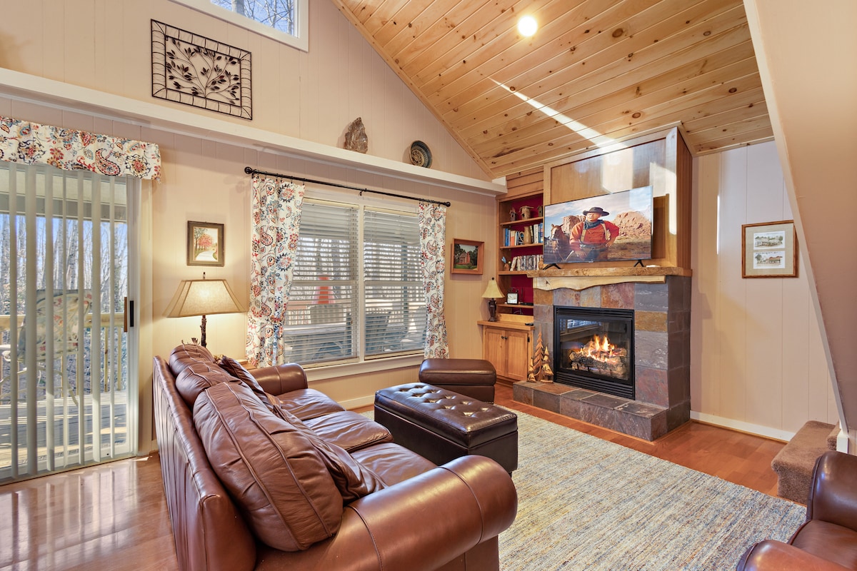 A cozy living area features a leather sofa and a coffee table on a textured rug. Natural light filters through patterned curtains, illuminating the space. A stone fireplace serves as a focal point, flanked by shelves with decorative items and books.