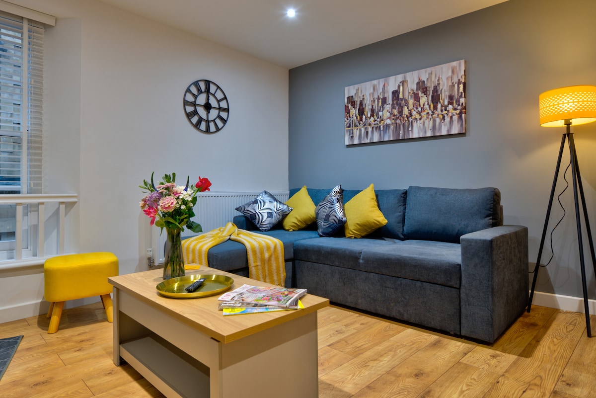 A modern living area presents a dark grey sofa adorned with vibrant yellow cushions, accompanied by a decorative throw. A coffee table holds a small vase of flowers and magazines, while a floor lamp adds subtle illumination. A large art piece depicting a cityscape hangs on the wall.