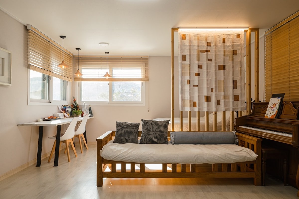 The living area features a light-colored sofa positioned beside a window, allowing natural light to fill the space. A dining table with white chairs is visible, along with decorative wooden partitions and warm pendant lighting overhead.