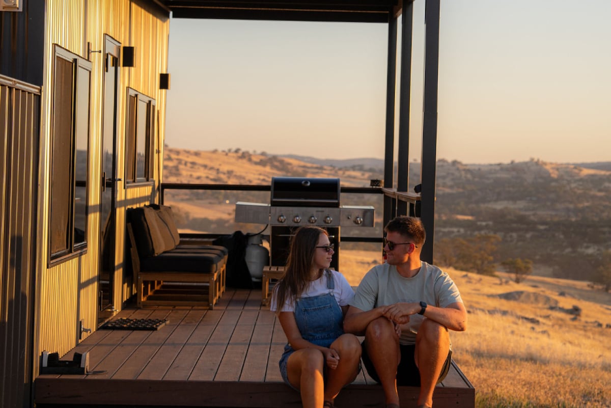 A spacious outdoor deck features a barbecue grill and comfortable seating, providing a perfect spot to enjoy sunsets over the surrounding hills. The landscape is characterized by rolling hills and warm tones, enhancing the relaxing ambiance.