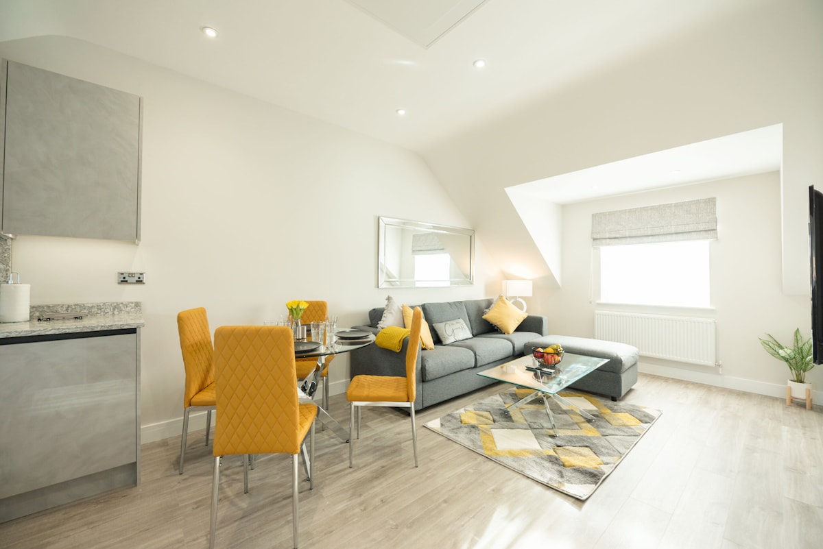 The open-plan living and kitchen area features modern furnishings with a comfortable sofa and a glass coffee table. A dining table with yellow upholstered chairs is set nearby. Light streams in from a window, illuminating the space, and a decorative rug adds warmth.