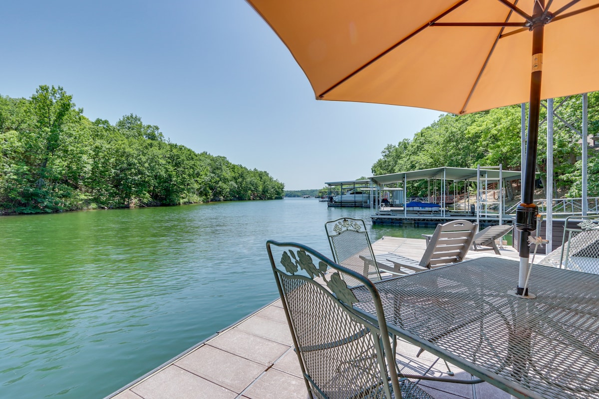 A shaded outdoor area features a large umbrella providing shade over a metal table with several chairs. The calm water reflects the adjacent greenery, creating a serene lakeside setting. Boat slips are visible along the shoreline, enhancing the aquatic atmosphere.