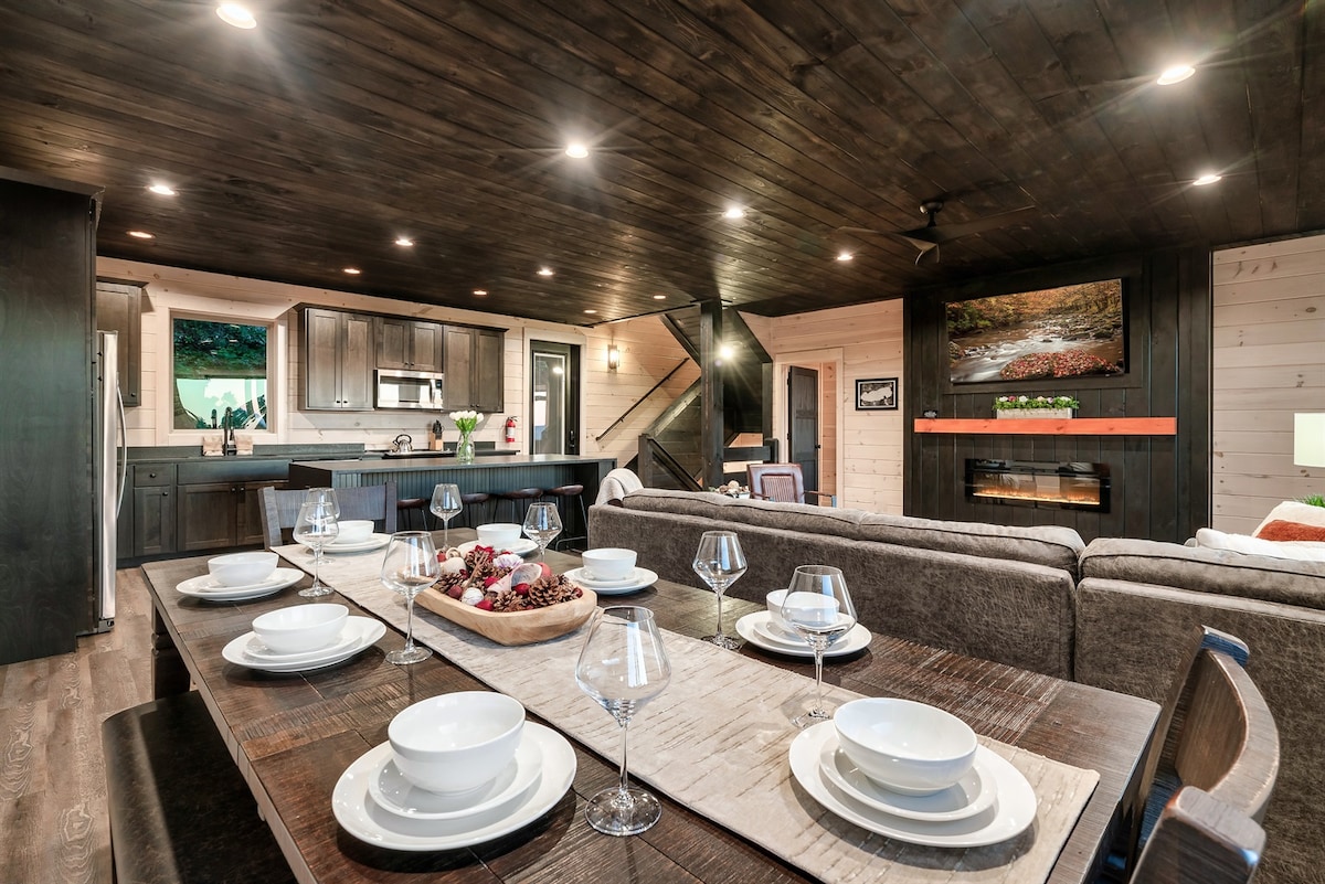 A spacious dining area is set with a large wooden table, accommodating multiple place settings of white plates and wine glasses. A cozy living room area with a sectional sofa is visible in the background, complemented by a modern kitchen featuring dark cabinetry and warm lighting.